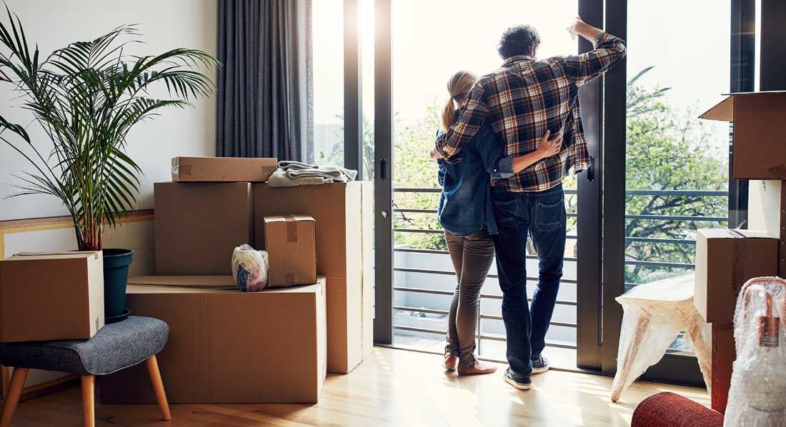 Ein Paar steht Arm in Arm am bodentiefen Fenster der neuen Wohnung und blickt hinaus, in der Wohnung stehen Umzugskartons und verpackte Möbel